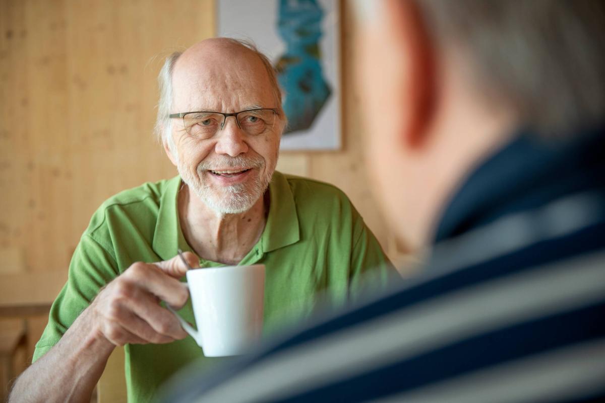 Vihreäpaitainen mies istumassa kahvikuppi kädessään. Etualalla näkyy toisen ihmisen hahmo takaapäin.